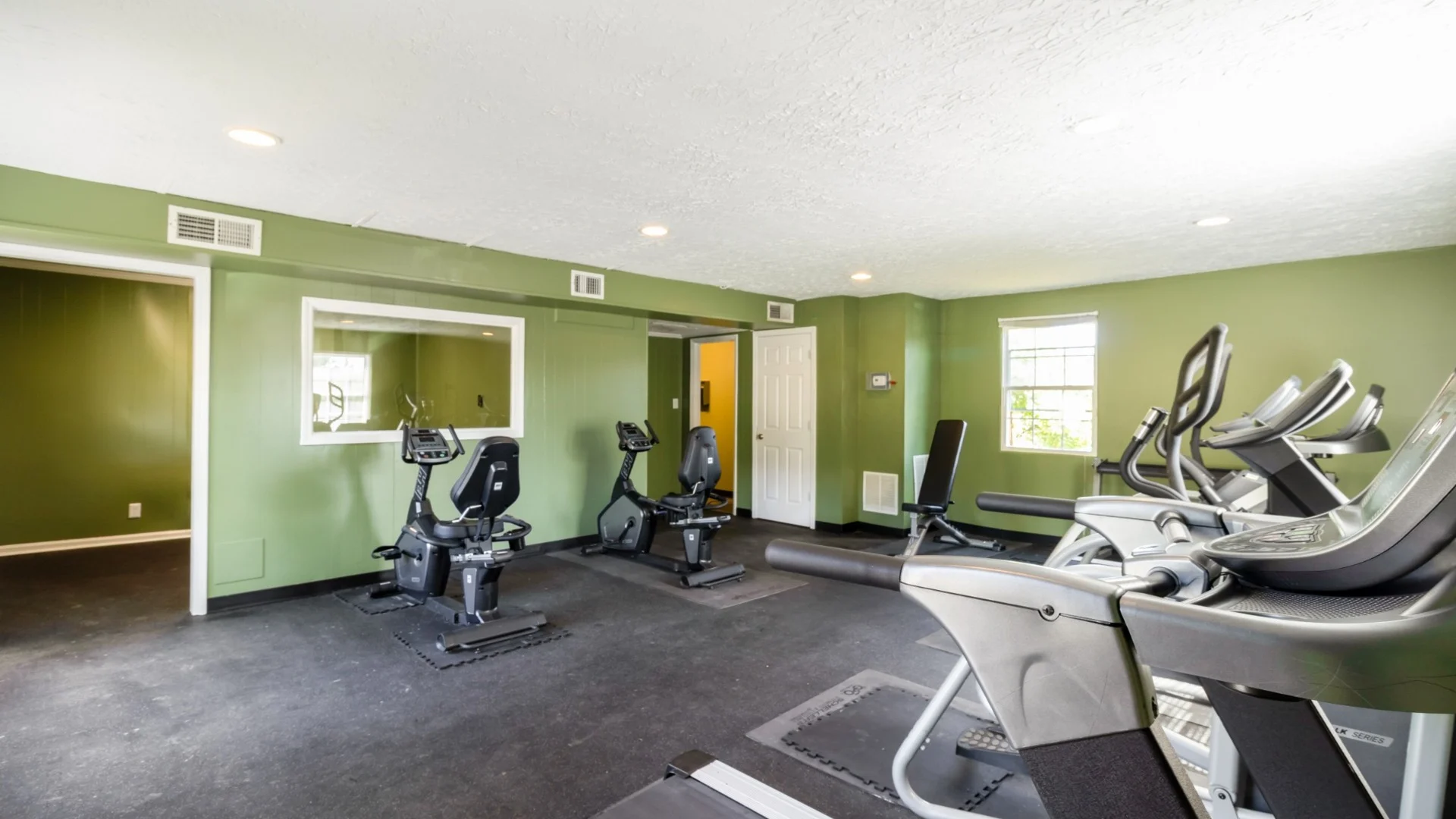 A well-lit indoor gym with exercise equipment, including stationary bikes and treadmills, set against green walls.