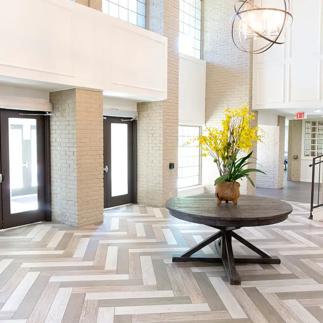 A spacious lobby with large windows and a stylish design featuring a round wooden table with a flower arrangement, patterned flooring, and double doors.