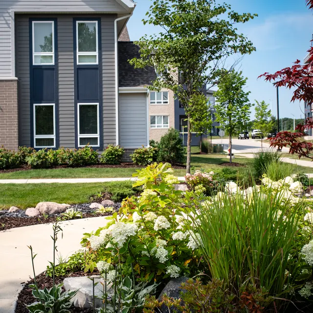 Modern Apartment Complex with Landscaped Gardens A modern apartment complex with landscaped gardens featuring green grass, flowers, and a walkway. Trees and shrubs are visible, and the sky is clear.