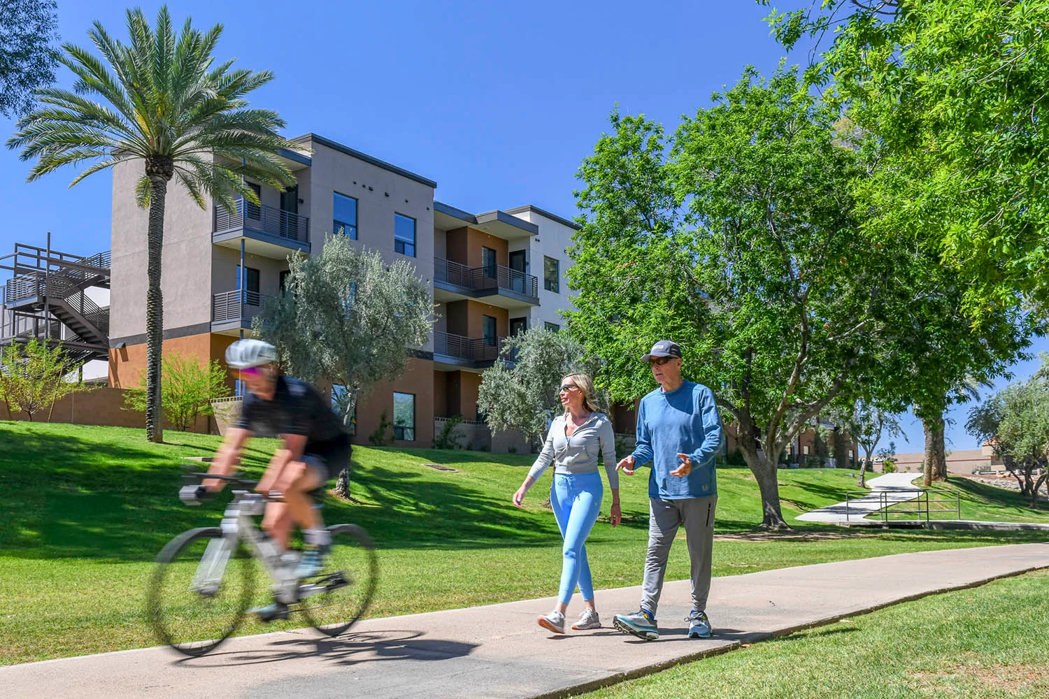 Couple Walking with Cyclist in Residential Area A cyclist rides past a couple walking on a pathway, surrounded by green grass and palm trees, with modern apartments in the background.