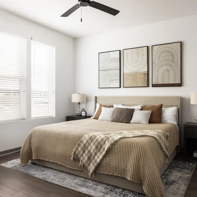 A modern bedroom featuring a king-size bed with a beige and cream blanket, two bedside lamps, and abstract wall art. Light streams in through large windows adorned with white blinds, accompanied by wooden flooring and a decorative rug.
