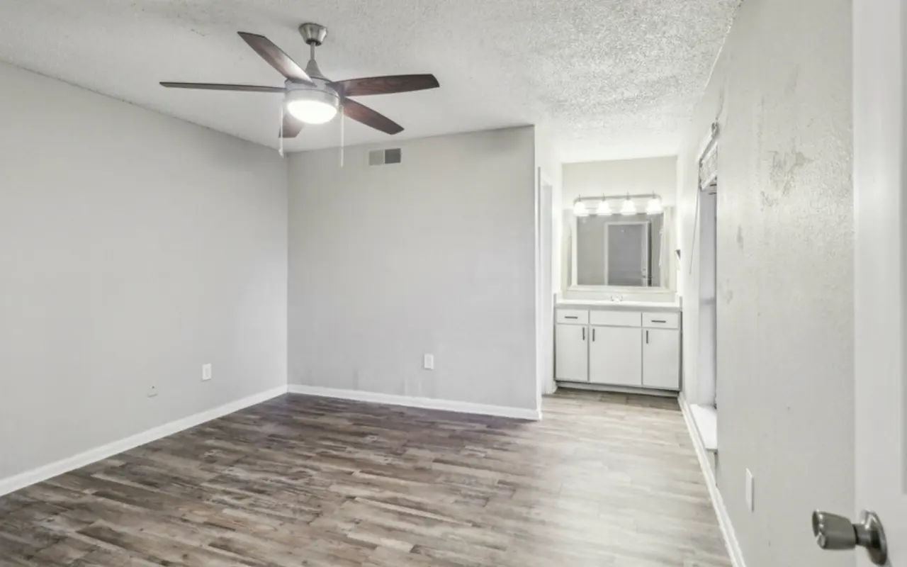 A spacious living room with a ceiling fan and wooden floor, leading to a vanity area with a mirror and sink.