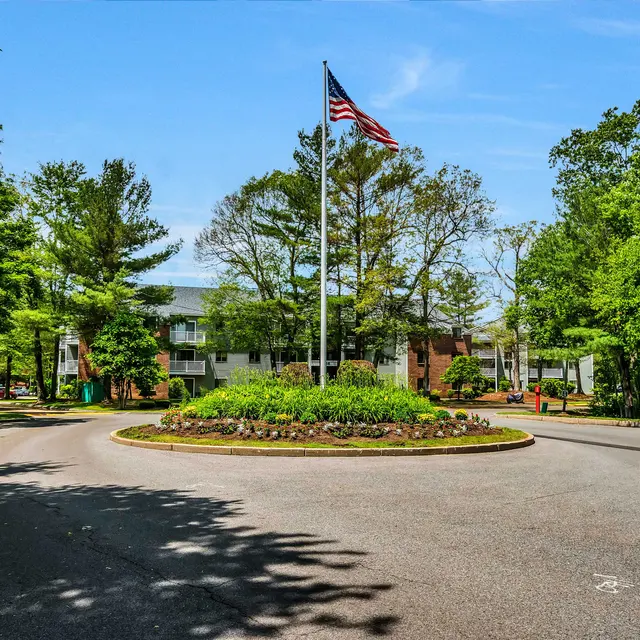 A circular driveway with a landscaped roundabout featuring a flagpole in the center, surrounded by trees and buildings in the background on a sunny day.