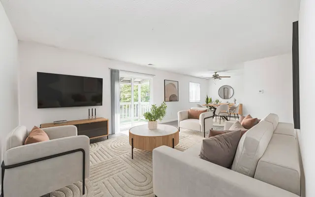 A modern living room featuring a large flat-screen TV, two light-colored sofas with decorative cushions, a round coffee table, and a dining area with a circular table and chairs. Large windows allow natural light in, and a ceiling fan is visible.