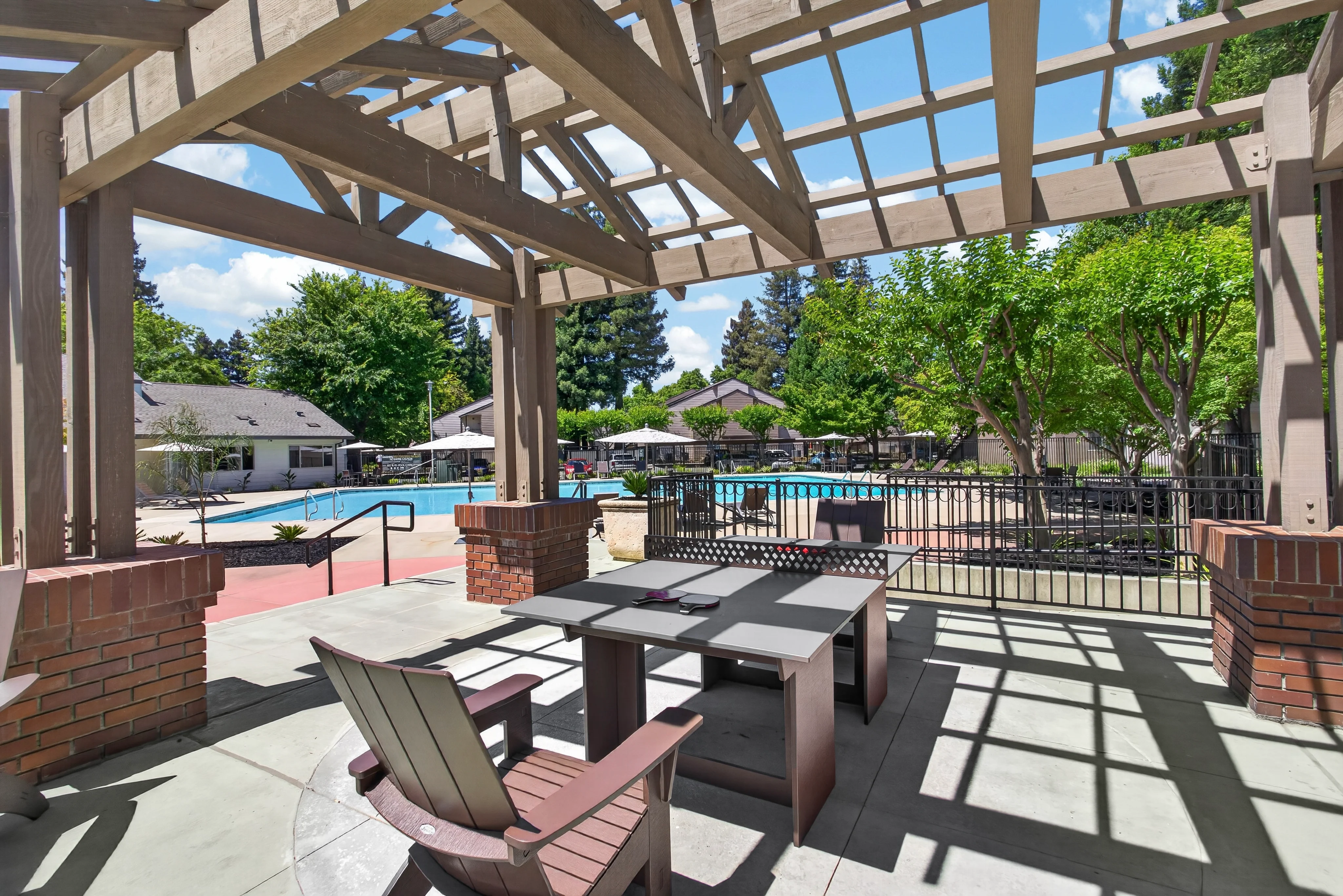 A patio area with a wooden structure overlooking a swimming pool. There are brown chairs and a grey table, surrounded by greenery and sunny skies.