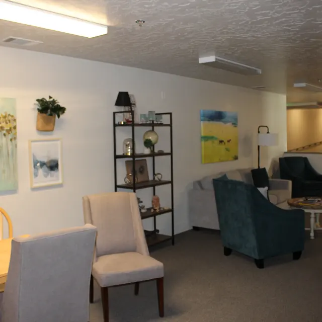 A spacious lounge area featuring a dining table with chairs, a small bookshelf, and comfortable seating arrangements. The walls are adorned with abstract artwork and there are plants for decoration.