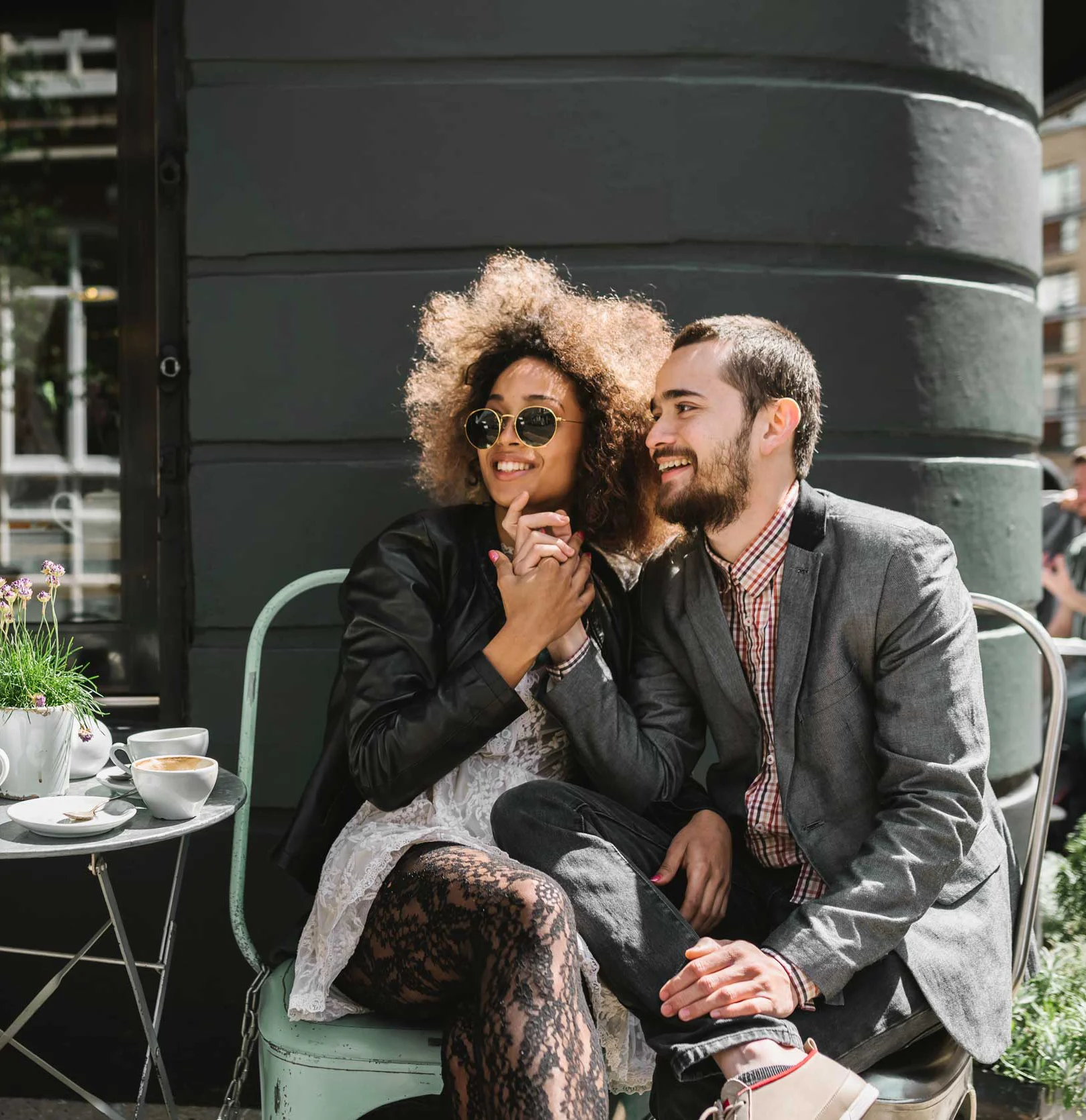 A couple sitting together at a café, the woman has curly hair and sunglasses, while the man is dressed in a blazer and smiling. They are holding hands and enjoying each other's company.