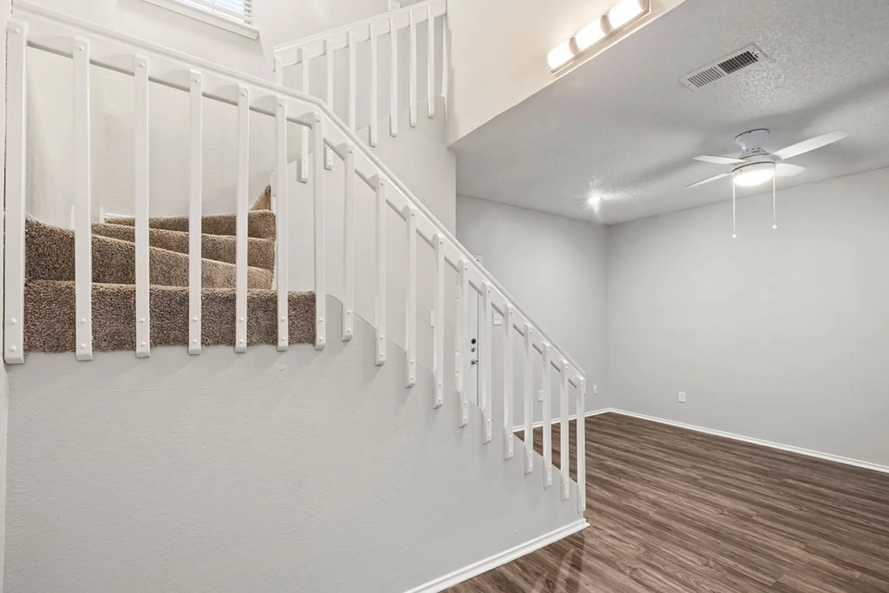 A view of a modern living space featuring a staircase with white railings and clean beige carpeting leading up. The floor is smooth and wooden, and there is a ceiling fan visible above. The walls are painted light gray, providing a neutral backdrop.
