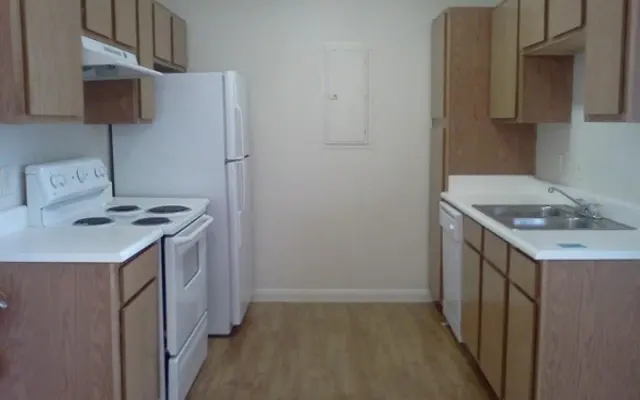 A simple kitchen featuring light-colored wooden cabinets, a white refrigerator, an oven, and a sink. The floor is a light wood laminate, and the walls are painted in a neutral color.