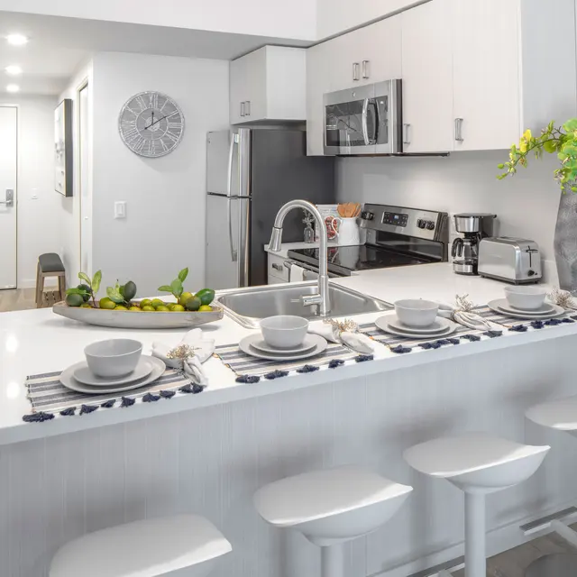 Modern Kitchen Interior A modern kitchen with a white countertop, bar stools, and stainless steel appliances. Decorative elements include a vase with green plants and a plate of fruits on the counter.