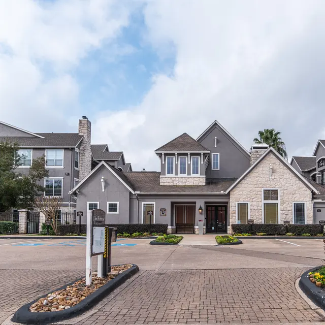 Front view of a modern apartment complex with gray walls and a stone entrance, surrounded by landscaping and cloudy sky.