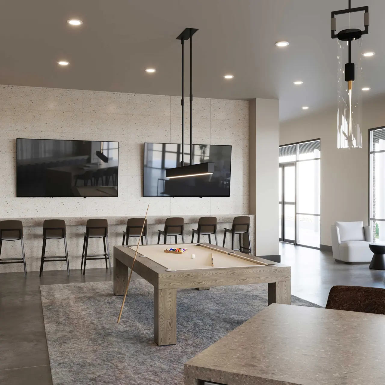 A modern game room featuring two pool tables, bar seating with high stools, and a pair of large wall-mounted televisions. The decor includes contemporary lighting fixtures and a neutral color palette with a rug on the floor. Windows let in natural light, and there are comfortable seating options in the corner.
