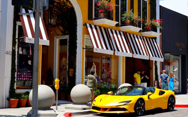 A vibrant yellow sports car parked alongside a trendy boutique with striped awnings, potted plants, and people walking by.