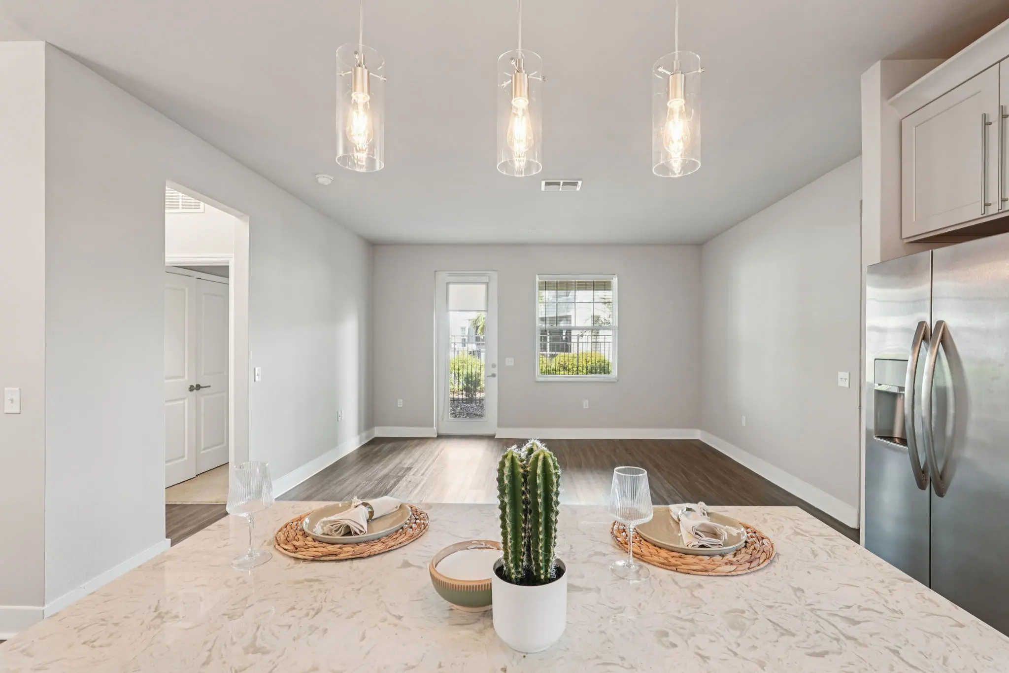 A modern kitchen island with cactus decor, two sets of plates, and a glass of water in the foreground. In the background, a spacious living area with large windows and recessed lighting is visible.