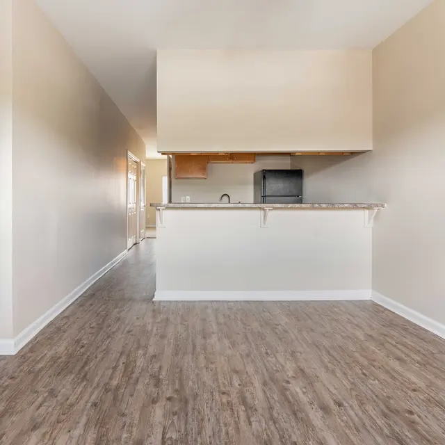 A spacious living area with wooden flooring leading to a small kitchen. A staircase is visible on the left, and the kitchen features a countertop and basic appliances.