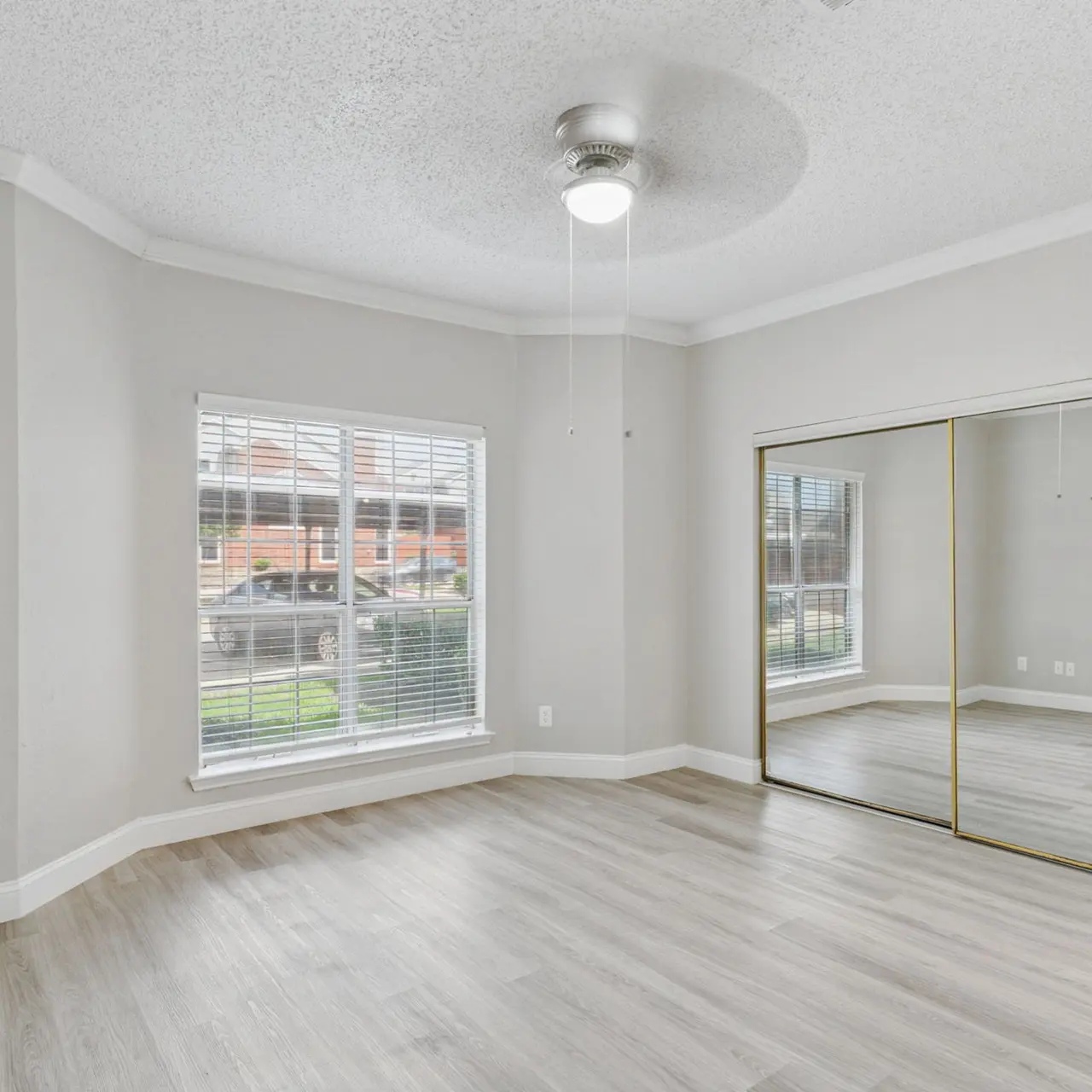 A bright and spacious bedroom featuring light-colored walls, wooden flooring, and a ceiling fan. There are large windows with blinds and a mirrored closet door.