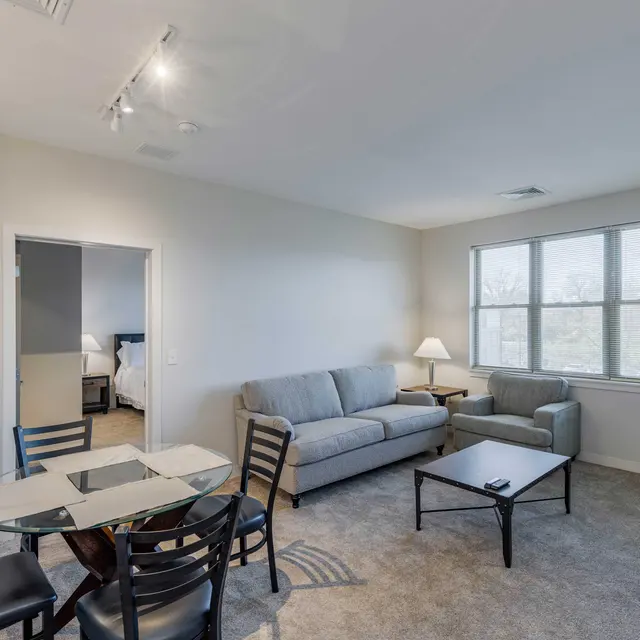 A cozy living room area featuring a gray sofa, a small armchair, a coffee table, and a glass dining table with black chairs. Large windows provide natural light, and a doorway leads to another room.