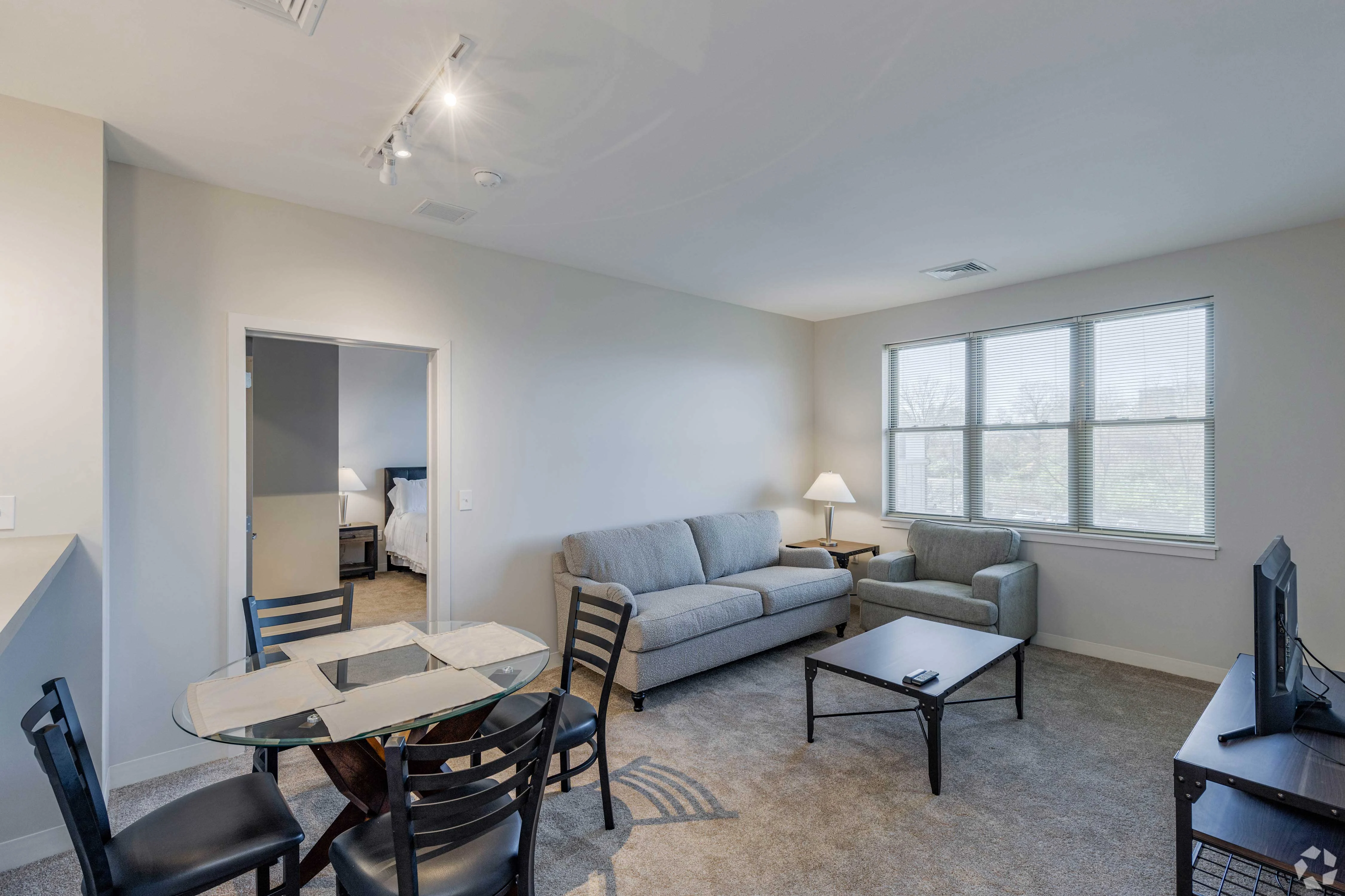 A cozy living room area featuring a gray sofa, a small armchair, a coffee table, and a glass dining table with black chairs. Large windows provide natural light, and a doorway leads to another room.