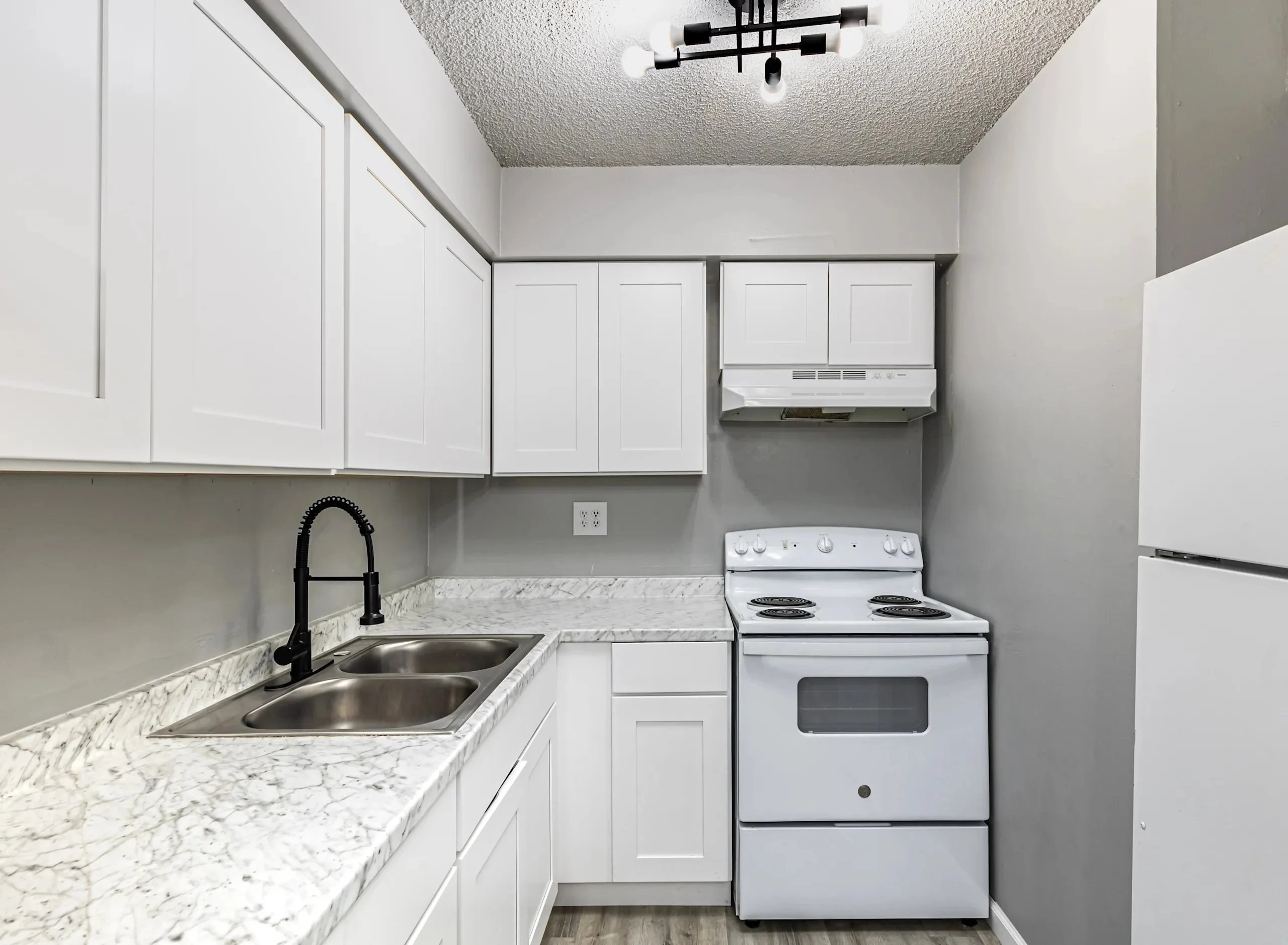 A modern kitchen with white cabinets, a marble countertop, a stainless steel sink, and a white stove under a ceiling light fixture.