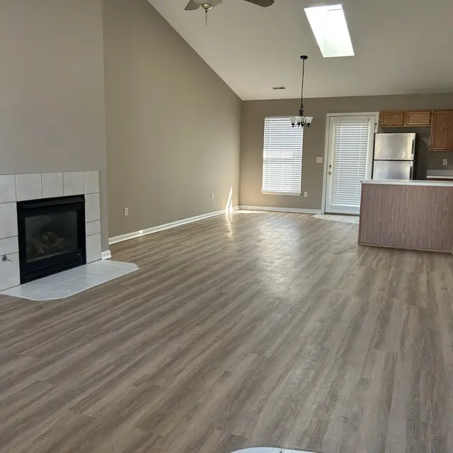 A spacious living room with high ceilings, a ceiling fan, and wooden flooring. There is a fireplace on the left side and an open kitchen area in the background, featuring light-colored cabinets and stainless steel appliances. Natural light enters through a skylight and a window.