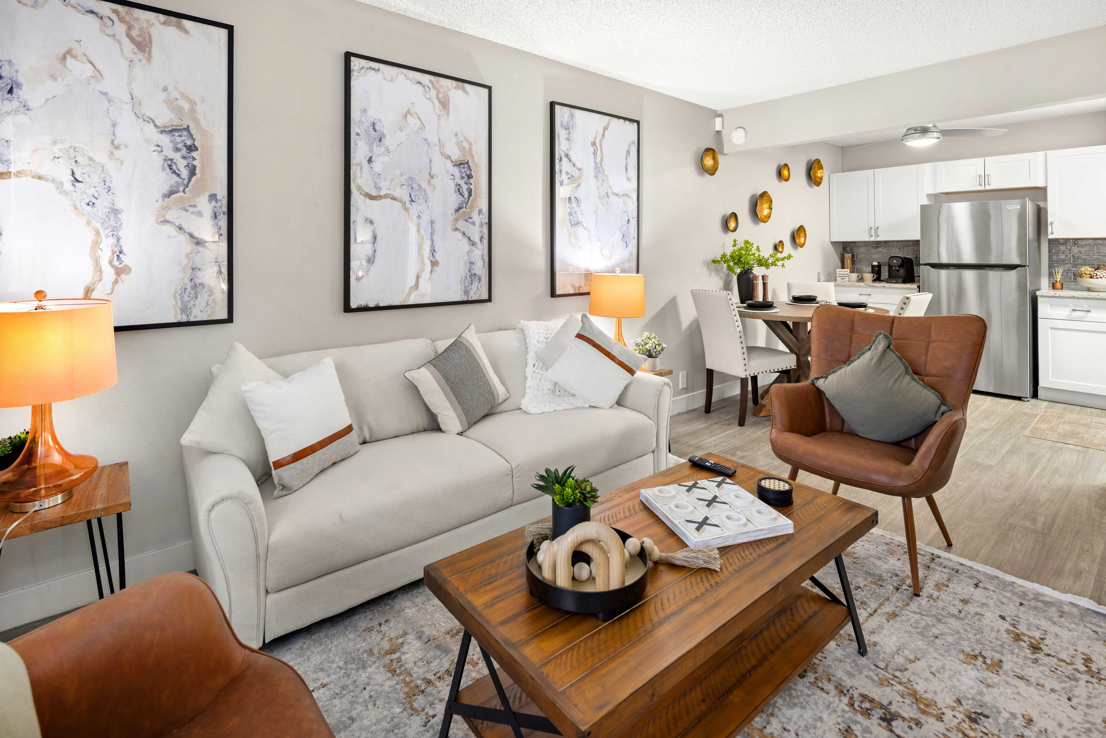 A modern living room featuring a light-colored sofa with pillows, a wooden coffee table, and stylish artwork on the walls. There are two lamps providing warm light, and a dining area is visible in the background.