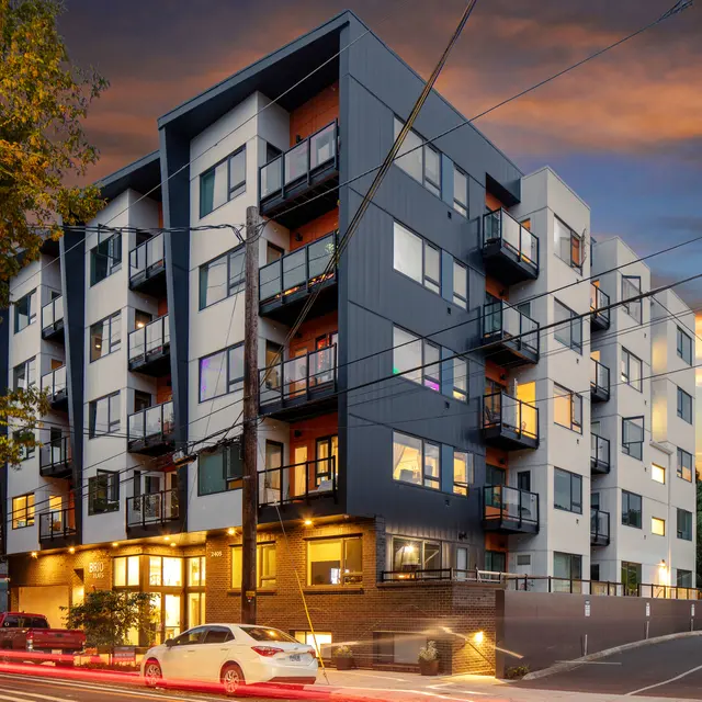Modern Apartment Building at Sunset A modern multi-story apartment building with balconies, illuminated windows, and a sunset sky. A white car is parked near the building.
