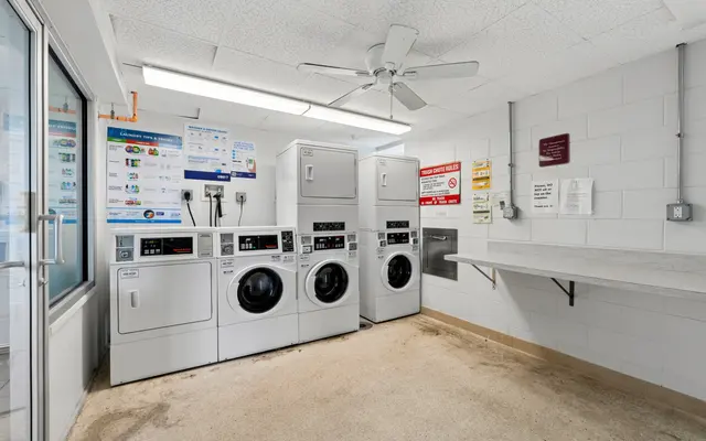 A clean laundry room featuring several washing machines, dryers, and a folding table. The walls are white with informational posters and signs.