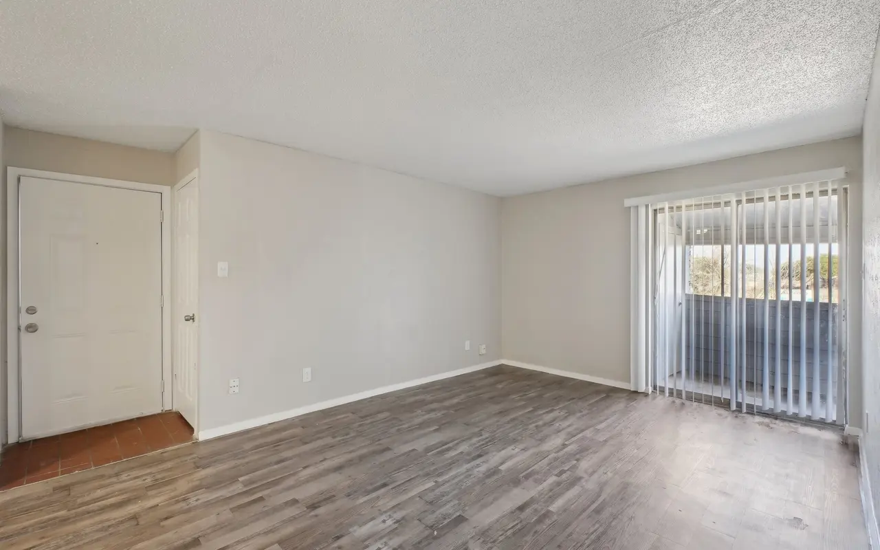 An empty living room with light-colored walls and hardwood-like flooring. There is a door on the left leading outside and a sliding glass door on the right that opens to a balcony or patio.