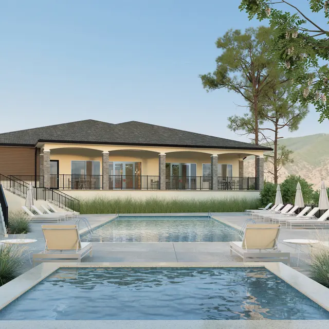 A modern pool area featuring a large house with a porch in the background, surrounded by green trees and mountains. There are lounge chairs arranged around the pool and a small water feature in the foreground.