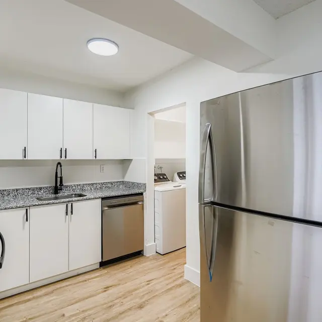 A modern kitchen featuring white cabinetry, a granite countertop, stainless steel appliances, and wood flooring. A doorway leads to a laundry area.