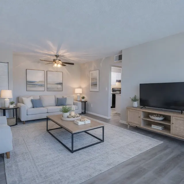 A modern living room featuring a light grey sofa, a wooden coffee table, a TV on a stand, and decorative plants. The walls are light-colored, adorned with artwork, and two lamps light the space.