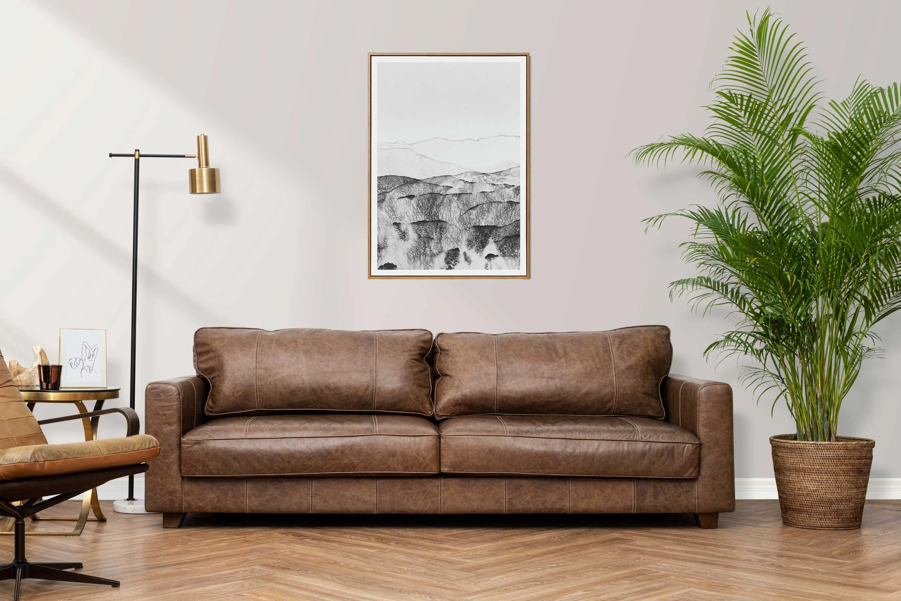 Stylish Living Room with Brown Leather Sofa A cozy living room featuring a brown leather sofa, a decorative wall art piece, a floor lamp, and a potted plant near a wooden floor.