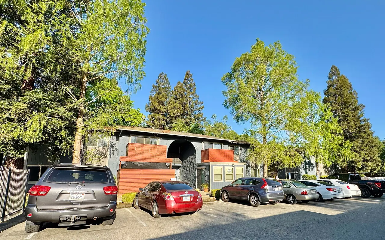 A view of a parking lot in front of an apartment complex with several parked cars and green trees in the background.