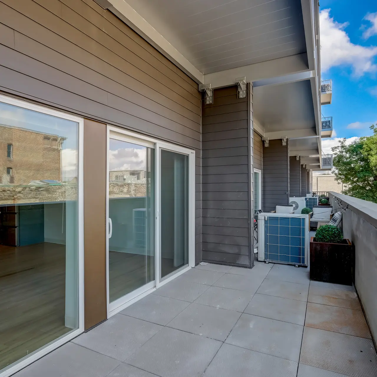 Element Apartments A modern balcony with sliding glass doors, featuring light-colored tile flooring, a small outdoor unit, and green plants against a backdrop of blue skies and clouds.