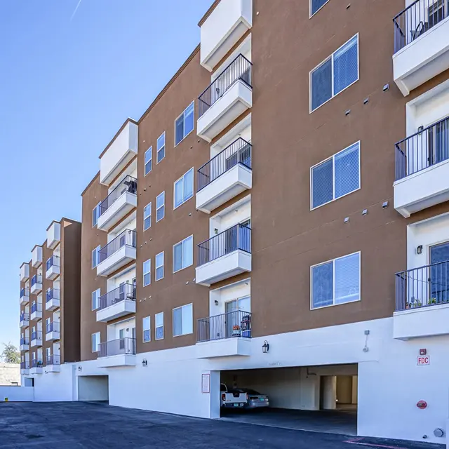 A modern apartment building with multiple floors, featuring balconies and large windows, set against a clear blue sky. The ground floor has parking beneath the building.