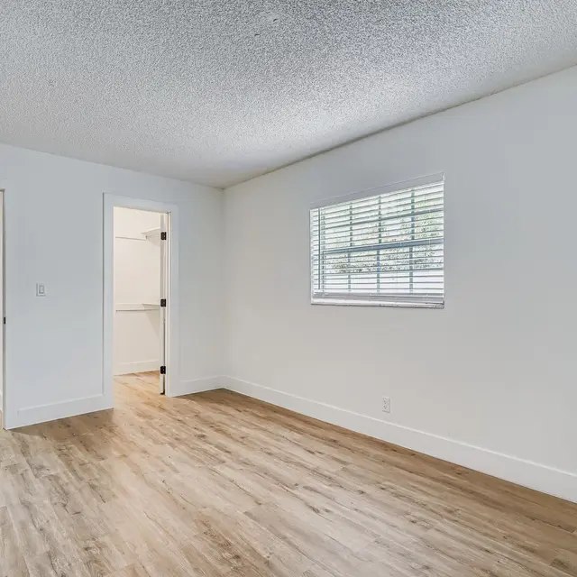 A spacious, empty room with light-colored walls and wooden flooring. There are two doorways visible, one leading to another room and the other to a hallway. A window with white blinds allows natural light to enter from the side. The overall atmosphere is bright and minimalistic.
