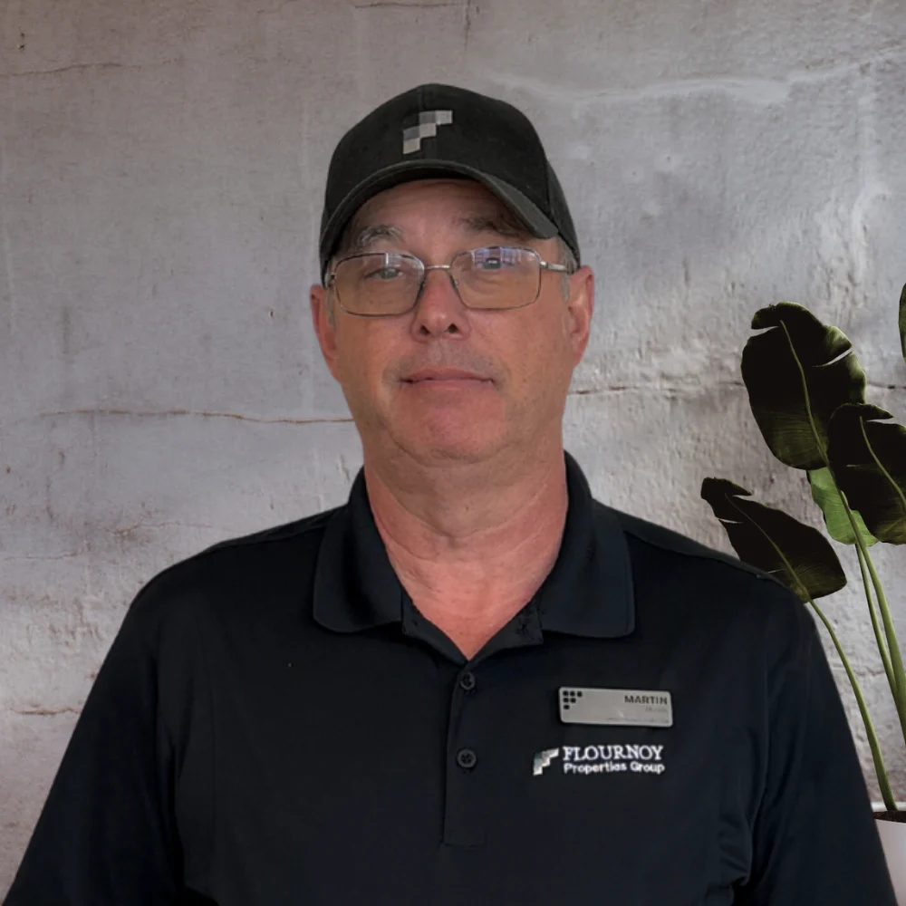 A man wearing a black shirt and a cap, standing against a textured wall with greenery in the background.