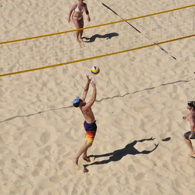 Aerial view of a beach volleyball game with players on sandy court. One player jumps to spike the ball while another player prepares to react. Two additional players can be seen in the background, and the volleyball net is visible.