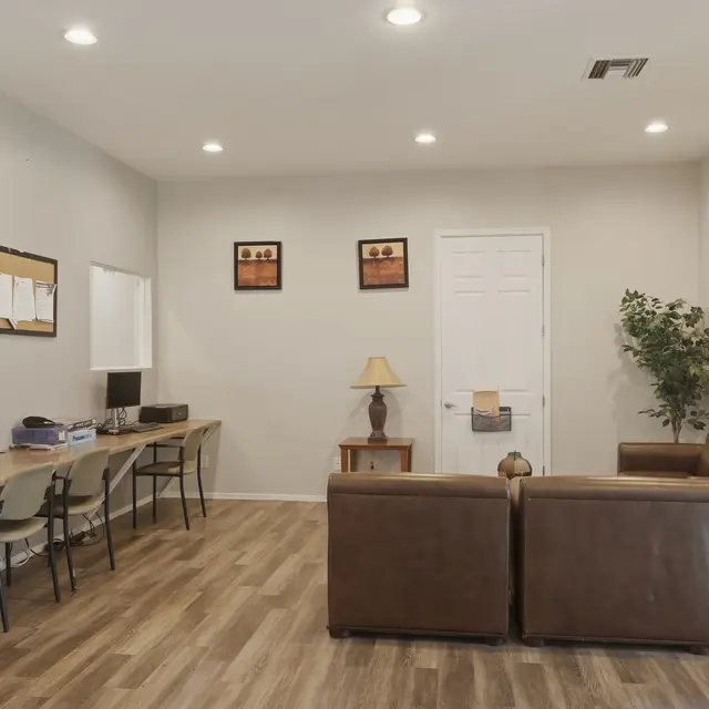 A modern waiting area with two brown couches, a lamp, and a small dining table with chairs. There are computers on a long desk and framed artwork on the walls.