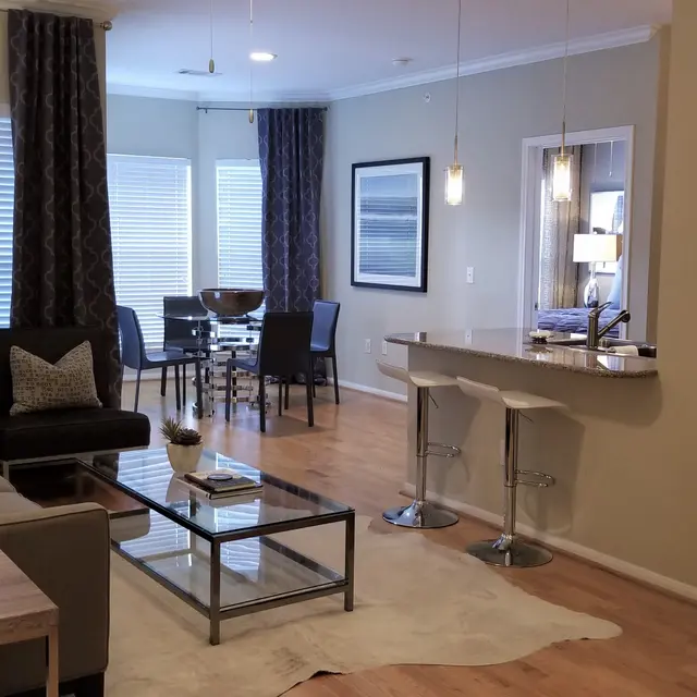 A modern living room featuring a comfortable grey sofa, a glass coffee table, and wooden accents. The space includes a dining area with a round table and chairs, with light streaming in from large windows. A flat-screen TV is mounted on the wall, and a stylish lamp adds warmth to the room.