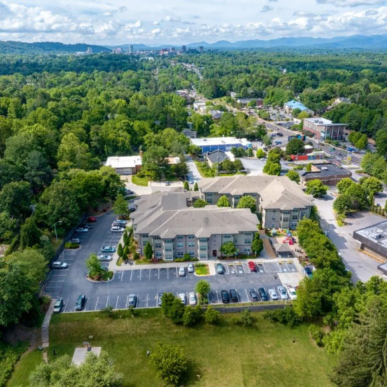 Aerial view of a residential area featuring an apartment complex surrounded by greenery and commercial buildings in the distance.