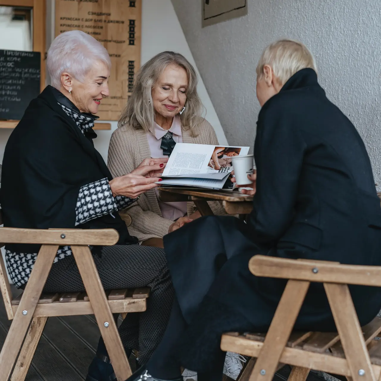 Three older women sitting together at a table, looking at a photo book and enjoying coffee in a casual setting.