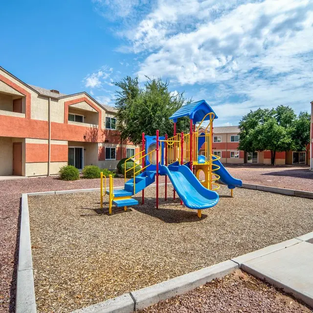 A colorful playground with slides and climbing structures surrounded by gravel, located in a courtyard of low-rise apartment buildings.