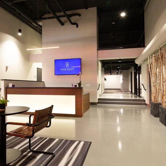 A modern lobby reception area featuring a wooden front desk, a circular table with chairs, a striped rug, and decorative wooden structures resembling trees along the wall.