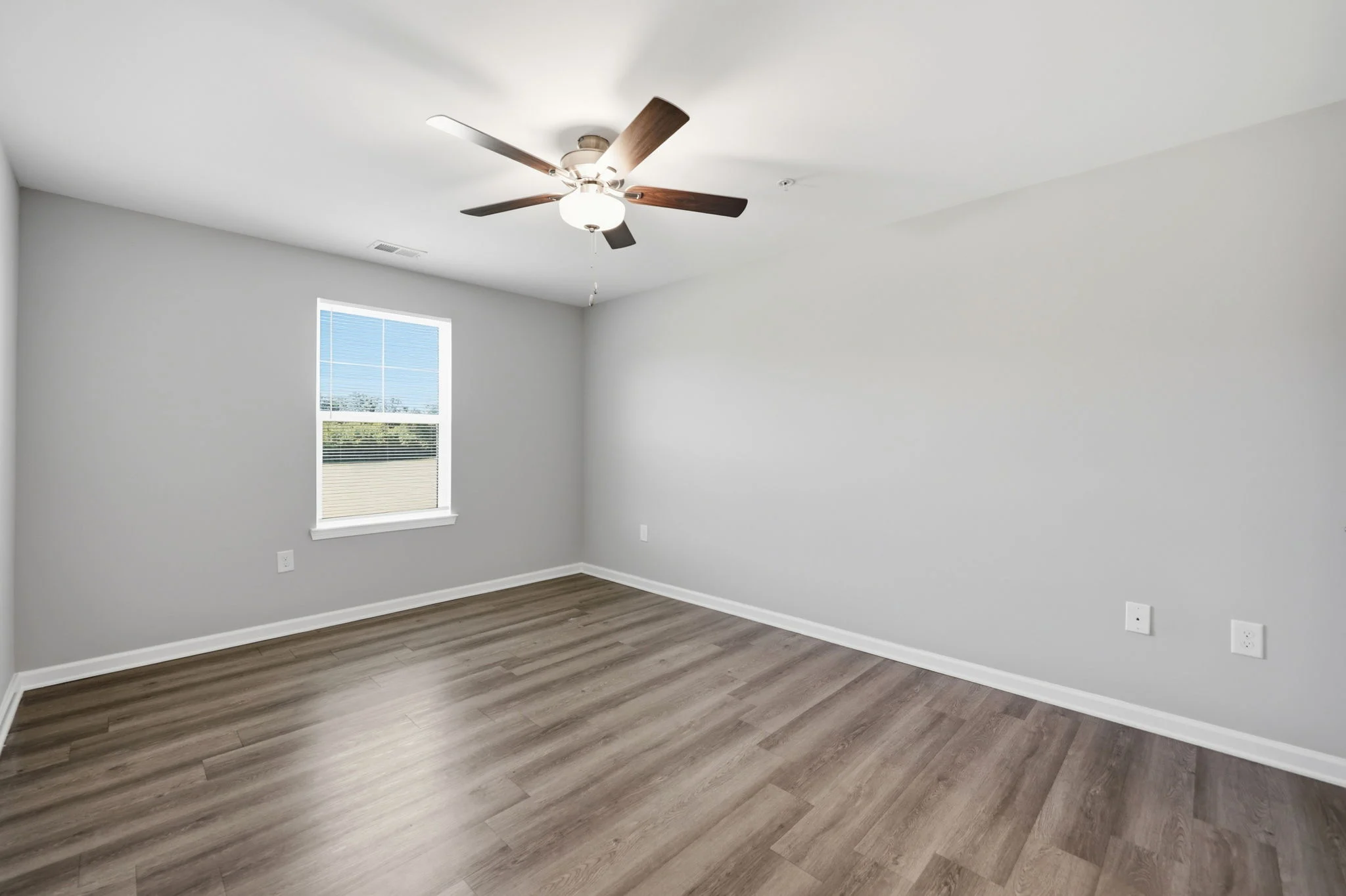 Empty Room with Ceiling Fan A spacious, empty room with gray walls and wooden flooring, featuring a ceiling fan and a window that lets in natural light.