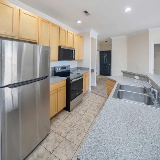 A modern kitchen featuring stainless steel appliances, wooden cabinets, and a granite countertop. There is an oven, microwave, and a large refrigerator. The flooring is tiled and the space is well-lit.