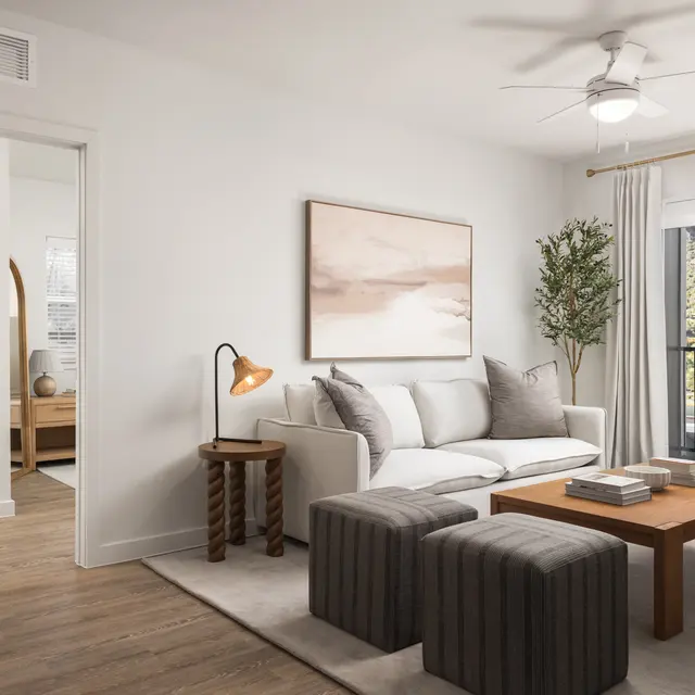 A modern living room with white walls, a comfortable couch, decorative pillows, a coffee table, and two ottomans. There is a lamp on a side table, and a large window offers a view of greenery outside.