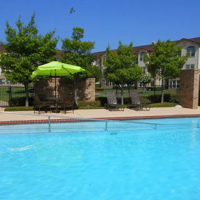 View of a swimming pool surrounded by green trees and lounge chairs under a bright blue sky.