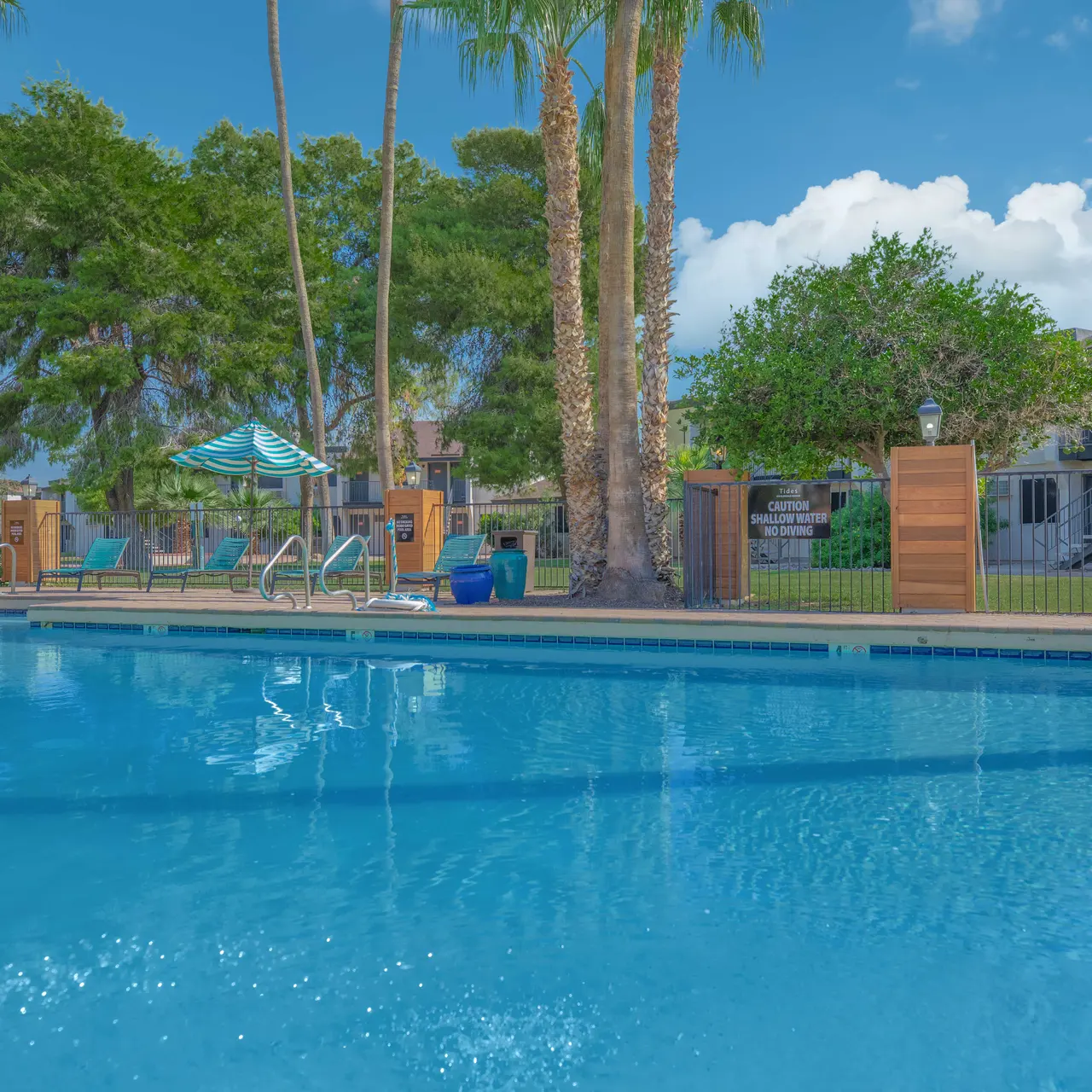 Swimming pool surrounded by palm trees and lounge chairs, with an apartment building in the background under a blue sky with clouds.