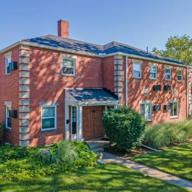 A two-story brick apartment building with a well-maintained lawn. The building features multiple windows and a front entrance with greenery around it.